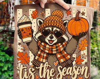 May include: A brown t-shirt featuring a graphic of a raccoon wearing a checkered beanie and scarf with heart-shaped sunglasses. The raccoon holds a coffee cup and a pumpkin. The text 'tis the season' is printed below with autumn leaves.