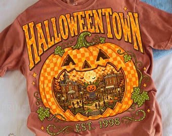 May include: A rust-colored t-shirt featuring a large graphic of a checkered pumpkin with a town scene inside. The text HALLOWEENTOWN is arched above in yellow and orange, with EST. 1998 printed below the pumpkin, surrounded by green vines.