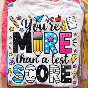 Puede incluir: Camiseta blanca con el mensaje "You're MORE than a test SCORE" en un dise&ntilde;o colorido y juguet&oacute;n. Presenta gr&aacute;ficos de l&aacute;pices, estrellas y tem&aacute;ticos escolares. La camiseta est&aacute; doblada y apilada con otras prendas.