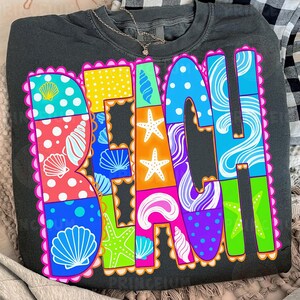 May include: Dark gray sweatshirt with the word "BEACH" in colorful, patterned letters. The letters are filled with images of seashells, starfish, and polka dots, outlined in pink. A silver necklace with a small pendant is visible.