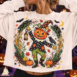 Può includere: Una felpa bianca a maniche lunghe con la grafica di uno spaventapasseri con testa di zucca, che indossa un cappello a scacchi, una giacca a pois e pantaloni verdi. Il disegno include una lanterna, un corvo nero, mais e foglie autunnali.