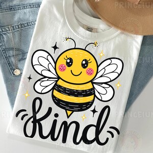 May include: White t-shirt featuring a cartoon bee with a yellow body, black stripes, and white wings. The word "Kind" is written in black script below the bee. The shirt is paired with blue jeans and a necklace.