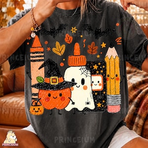 Op de afbeelding: Een antracietgrijs t-shirt met een grafische print van een lachende oranje pompoen met een heksenhoed, een witte geest met een spinnenweb, een kleurpotlood, een potlood en een zwart notitieboek, omringd door kleine oranje sterren en herfstbladeren.