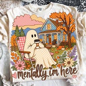 May include: A cream-coloured t-shirt featuring a graphic of a ghost sitting in a pink checkered lawn chair, holding a coffee cup. The scene includes a blue house, autumn trees, pumpkins, and flowers. The text "mentally im here" is printed below.