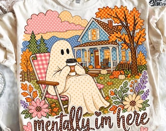May include: A cream-colored t-shirt featuring a graphic of a ghost sitting in a pink checkered lawn chair, holding a coffee cup. The scene includes a blue house, autumn trees, pumpkins, and flowers. The text "mentally im here" is printed below.