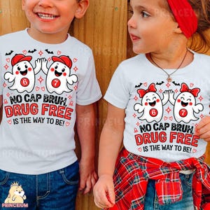 May include: Two white t-shirts featuring a graphic of two cartoon ghosts wearing red caps and bows. The ghosts have the numbers 6 and 7 on their chests. The text reads: NO CAP BRUH, DRUG FREE IS THE WAY TO BE! The design includes small bats and hearts.