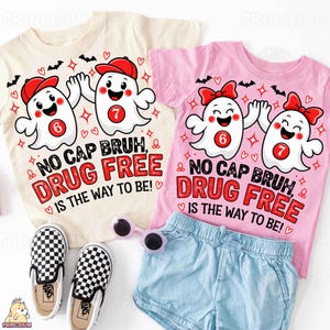 May include: A cream t-shirt and a pink t-shirt, both featuring a graphic of two ghosts with red caps or bows and the text NO CAP BRUH, DRUG FREE IS THE WAY TO BE, alongside a pair of light blue shorts and black and white checkered slip-on shoes.