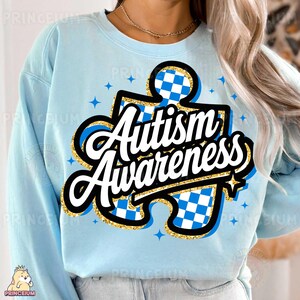 May include: Light blue sweatshirt featuring a gold glitter puzzle piece design. The puzzle piece has a blue and white checkered pattern and the text "Autism Awareness" in white.
