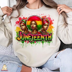K&ouml;nnte beinhalten: Ein wei&szlig;er Pullover mit einem farbenfrohen Grafikdesign, das drei Frauen mit buntem Haar und Kopfbedeckungen zeigt. Die Grafik enth&auml;lt auch das Wort "Juneteenth" in einer bunten, Graffiti-artigen Schrift.