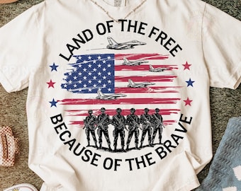 May include: White t-shirt with a red, white, and blue American flag design. The text "Land of the Free Because of the Brave" is printed around the flag. There are silhouettes of soldiers standing in front of the flag.