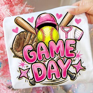 May include: White t-shirt with a softball design. It features a pink helmet, a yellow softball, a brown glove, and the words "GAME DAY" in pink bubble letters. Pink hearts and stars are also included in the design.