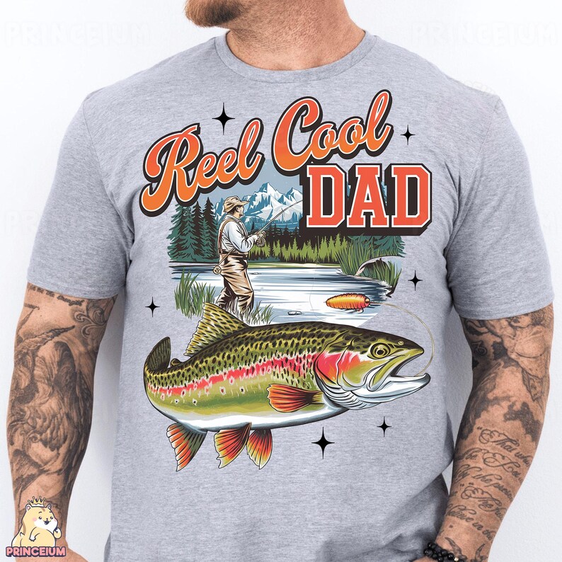May include: A light gray t-shirt featuring a colorful graphic of a trout fish and a fisherman. The text "Reel Cool Dad" is in orange and red. The design includes a lake and mountain scene.