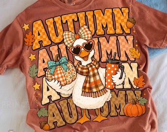 May include: A rust-colored t-shirt featuring a graphic of a duck wearing heart-shaped sunglasses and a plaid scarf. The duck holds a plaid pumpkin and a polka-dot tumbler. The text AUTUMN is repeated in a checkered font with autumn leaves.
