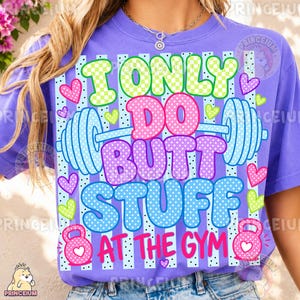 May include: A purple t-shirt featuring a graphic print with the text I ONLY DO BUTT STUFF AT THE GYM. The design includes a light blue barbell, two pink kettlebells, and various colorful hearts against a background of vertical stripes.