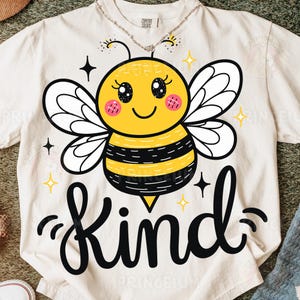 May include: Cream-colored t-shirt featuring a cartoon bee with a yellow body, black stripes, and white wings. The word "Kind" is written in black script below the bee, with star and sparkle accents. The shirt is a Comfort Colors brand.