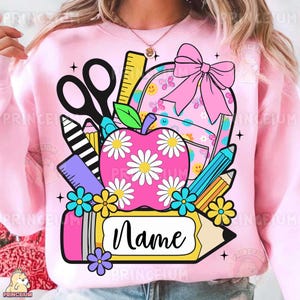 Könnte beinhalten: Rosa Sweatshirt mit einem Schul-Themen-Motiv. Das Design umfasst einen rosa Rucksack mit einer Schleife, einen Apfel mit Gänseblümchen, Bleistifte, eine Schere und ein Lineal. Ein gelber Bleistift mit Namensschild befindet sich unten.
