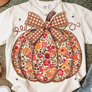 May include: Cream-colored t-shirt featuring a large pumpkin design. The pumpkin is filled with a floral pattern in shades of red, orange, and brown, with a brown plaid bow. The shirt has a Comfort Colors tag.