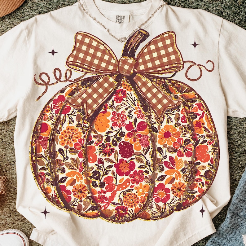 May include: Cream-colored t-shirt featuring a large pumpkin design. The pumpkin is filled with a floral pattern in shades of red, orange, and brown, with a brown plaid bow. The shirt has a Comfort Colors tag.