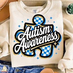 May include: A beige sweatshirt featuring a blue and white jigsaw puzzle design. The puzzle piece has gold glitter and the words "Autism Awareness" in white lettering.