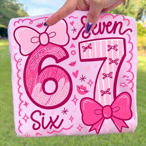 Może przedstawiać: Różowa koszulka z nadrukiem z cyframi "6" i "7" w zabawnym designie. Słowo "Six" jest napisane pod cyfrą 6. Słowo "Seven" jest napisane nad cyfrą 7. Projekt zawiera kokardy, serca i gwiazdy.