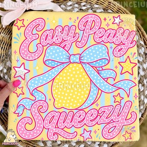 Puede incluir: Una camiseta amarilla con un gráfico central de un limón atado con un lazo azul claro de lunares. El texto "Easy Peasy Squeezy" está impreso en letras grandes de color rosa con lunares blancos. El fondo tiene rayas verticales amarillas y azules.
