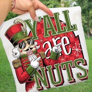 May include: White square pillow with a cartoon nutcracker design. The nutcracker wears sunglasses and a red and green uniform. Text reads "Y'ALL are NUTS" in green and white. The background has a red and white checkered pattern with snowflakes.