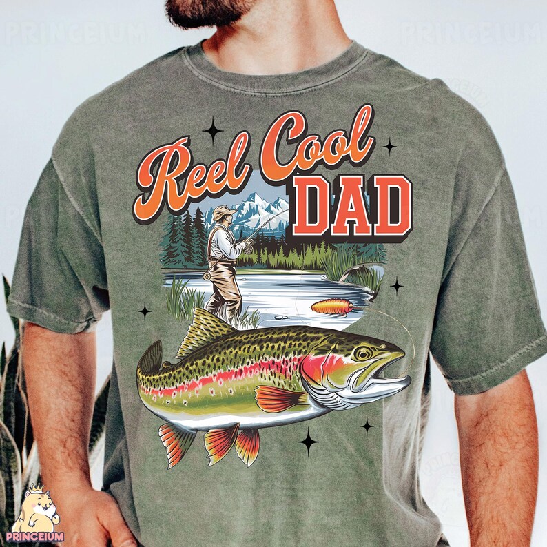 May include: Olive green t-shirt with a graphic featuring a fisherman, a trout, and the text "Reel Cool Dad" in orange and white. The design includes a mountain and lake scene, with a fishing lure.