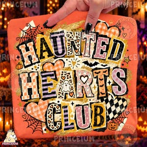 Op de afbeelding: Een oranje t-shirt met een grafische print met de tekst "HAUNTED HEARTS CLUB" in dikke, verweerde letters. Het ontwerp bevat patronen van geesten, luipaardvlekken, geblokte harten, spinnenwebben, vleermuizen en gouden glitteraccenten.