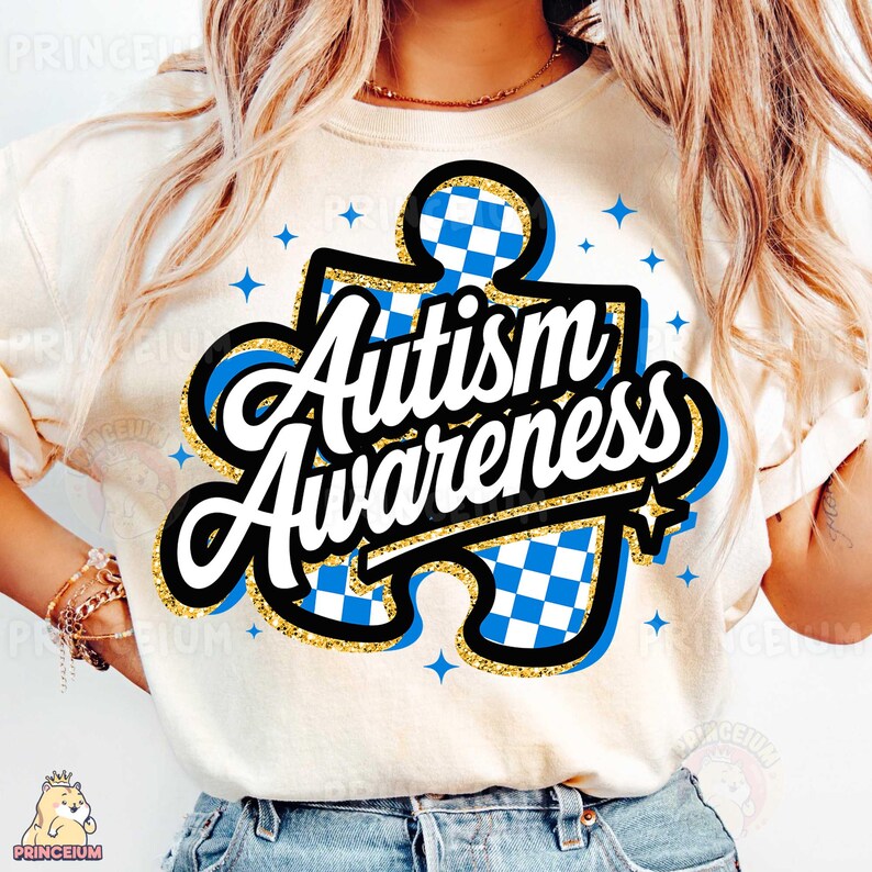 May include: A white t-shirt featuring a blue and white checkered puzzle piece design. The words "Autism Awareness" are written in a black and gold font over the puzzle piece.