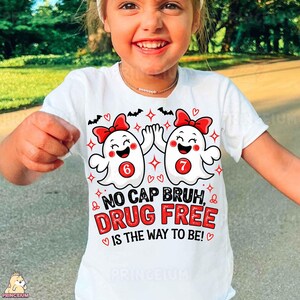 May include: A white short-sleeved t-shirt featuring a graphic of two white ghosts with red bows, each displaying a red circle with the numbers 6 and 7. The text reads NO CAP BRUH, DRUG FREE IS THE WAY TO BE! surrounded by small red hearts and black bats.