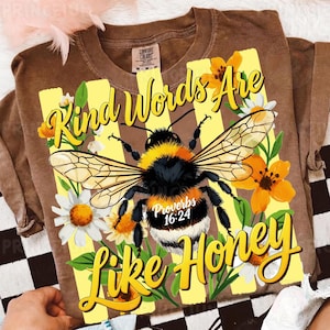 May include: Brown t-shirt with a large graphic of a bumblebee, flowers, and the text "Kind Words Are Like Honey" and "Proverbs 16:24". The design features yellow and white daisies and orange flowers.