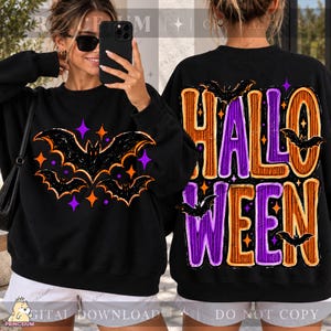 May include: A black long-sleeve sweatshirt featuring a graphic print of three black bats surrounded by orange and purple stars on the front. The back displays the large text HALLOWEEN in orange and purple with additional bat and star graphics.