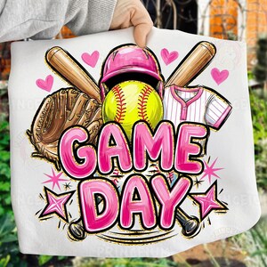 May include: A white design with a softball theme, including a yellow softball, pink helmet, baseball bat, glove, and jersey. The words "GAME DAY" are in pink with a gold outline, surrounded by pink hearts and stars.