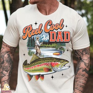 May include: Cream-colored t-shirt with a fishing-themed graphic. The design features the text "Reel Cool DAD" in orange and red, a fisherman, a lake, and a colorful fish. The shirt is a casual wear item.