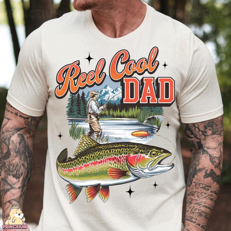 May include: Cream-colored t-shirt with a fishing-themed graphic. The design features the text "Reel Cool DAD" in orange and red, a fisherman, a lake, and a colorful fish. The shirt is a casual wear item.