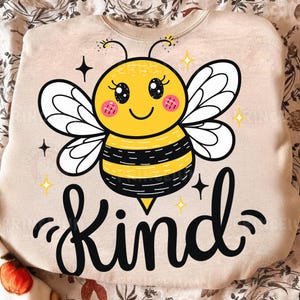 May include: A beige sweatshirt featuring a cartoon bee with a yellow body, black stripes, and white wings. The bee has a smiling face and the word "Kind" in black script below. The design includes stars and sparkles.