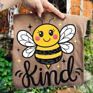 May include: A brown square pillow featuring a cartoon bee with a yellow body, black stripes, and white wings. The bee has a smiling face and pink cheeks. The word "Kind" is written in black script below the bee, with star accents.