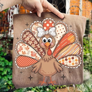 Könnte beinhalten: Ein braunes quadratisches Kissen mit einer Cartoon-Truthahn mit einer weißen Schleife und gemusterten Federn in Orange-, Braun- und Cremetönen. Der Truthahn hat einen roten Wamm und ist von Sternakzenten umgeben. Das Kissen wird von einer Hand gehalten.