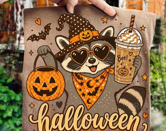May include: A brown t-shirt featuring a graphic of a raccoon wearing a polka-dot witch hat with a pumpkin, heart-shaped sunglasses, and an orange bandana. The raccoon holds a jack-o'-lantern bucket and a coffee cup with the text Boo! and halloween.