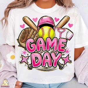 May include: White t-shirt with a softball design. The graphic includes a yellow softball, pink helmet, baseball bat, glove, and jersey. The words "GAME DAY" are in large, pink bubble letters, with star accents.