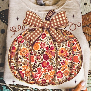 May include: A light beige sweatshirt featuring a large pumpkin design. The pumpkin is filled with a floral pattern in shades of red, orange, and brown, with a brown plaid bow. The design is outlined in gold glitter.