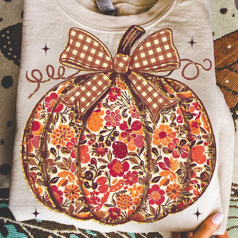 May include: A light beige sweatshirt featuring a large pumpkin design. The pumpkin is filled with a floral pattern in shades of red, orange, and brown, with a brown plaid bow. The design is outlined in gold glitter.