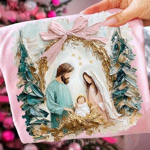May include: A light pink t-shirt featuring a textured, impasto-style oil painting print of a nativity scene. The design depicts a central figure in a teal tunic, a figure in a white veil, and a swaddled infant, framed by a large pink bow and pine trees.