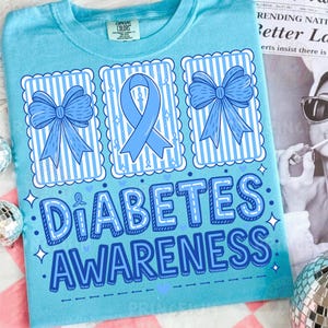 May include: Light blue t-shirt with the words "DIABETES AWARENESS" in a bold, outlined font. The design includes three framed images with blue bows and a blue ribbon. The shirt is a comfortable, casual wear item.