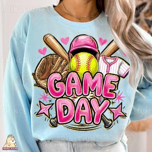 May include: Pale blue sweatshirt with a softball design. The graphic includes a yellow softball, pink helmet, baseball bat, glove, and the words "GAME DAY" in pink. The design also features pink hearts and stars.