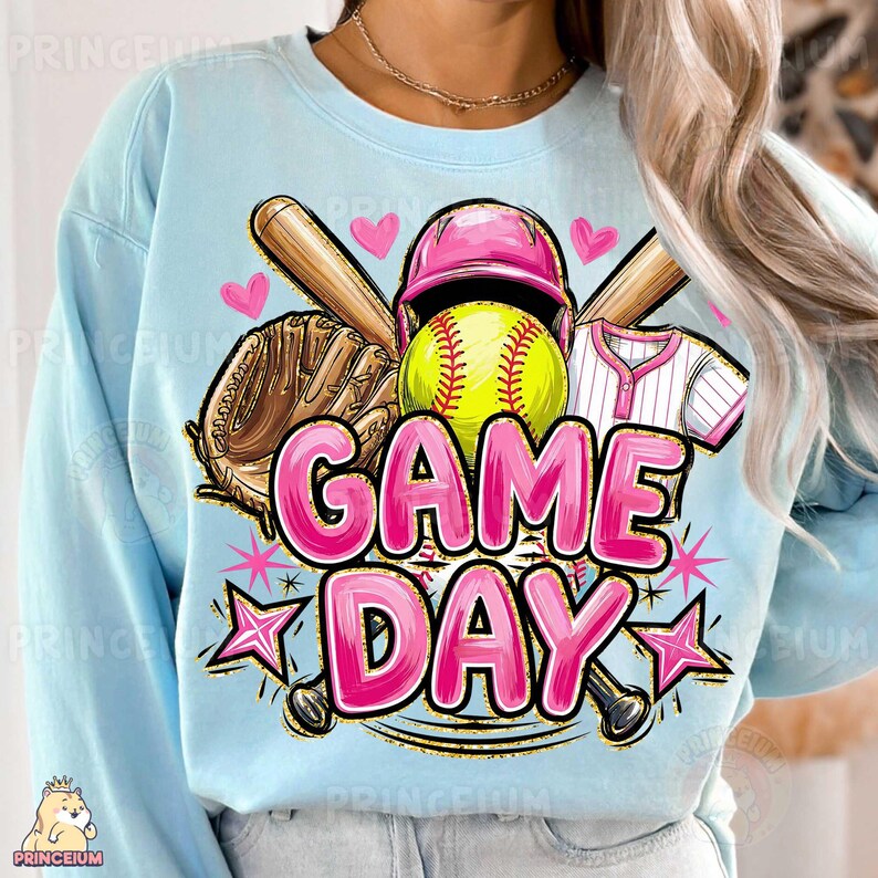 May include: Pale blue sweatshirt with a softball design. The graphic includes a yellow softball, pink helmet, baseball bat, glove, and the words "GAME DAY" in pink. The design also features pink hearts and stars.