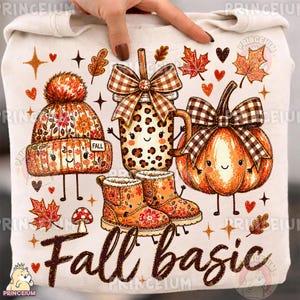 May include: A cream-coloured sweatshirt featuring a graphic print of a winter hat, a leopard-print mug with a plaid bow, a pumpkin with a plaid bow, and a pair of boots, all with faces. The text "Fall basic" is printed in brown script below.