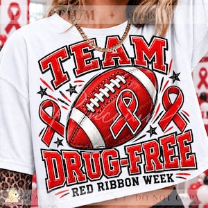 May include: A white t-shirt featuring a graphic of a red and white football centered between two red awareness ribbons. The text reads "TEAM DRUG-FREE" in bold red letters with black outlines, and "RED RIBBON WEEK" in smaller black text below the ball.
