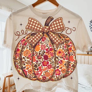 May include: Cream-colored t-shirt featuring a large pumpkin design. The pumpkin is filled with colorful floral patterns in shades of orange, red, and yellow. A brown plaid bow sits atop the pumpkin, with decorative swirls and stars around the design.