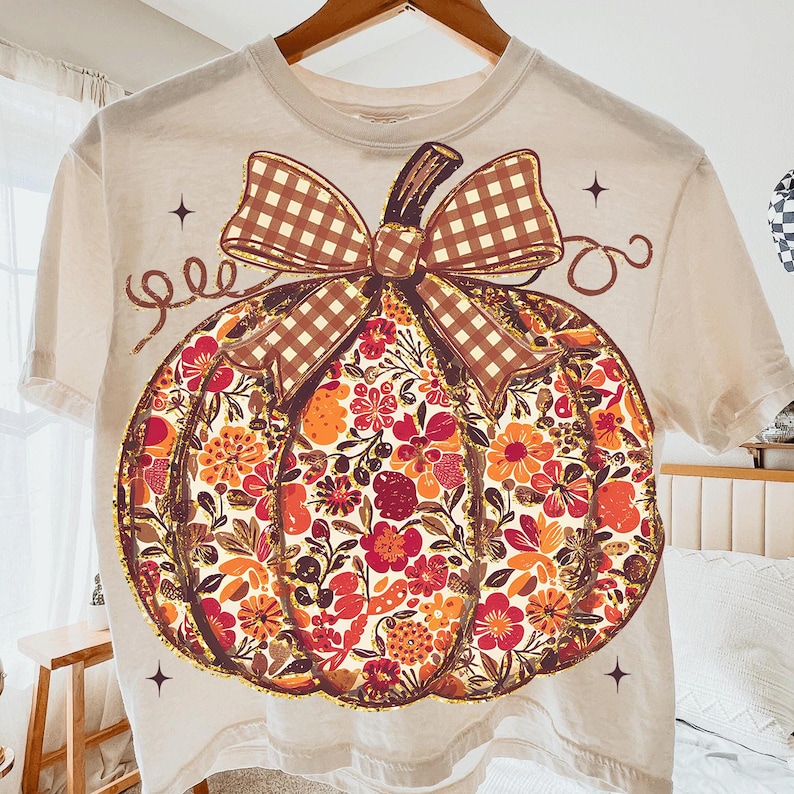 May include: Cream-colored t-shirt featuring a large pumpkin design. The pumpkin is filled with colorful floral patterns in shades of orange, red, and yellow. A brown plaid bow sits atop the pumpkin, with decorative swirls and stars around the design.